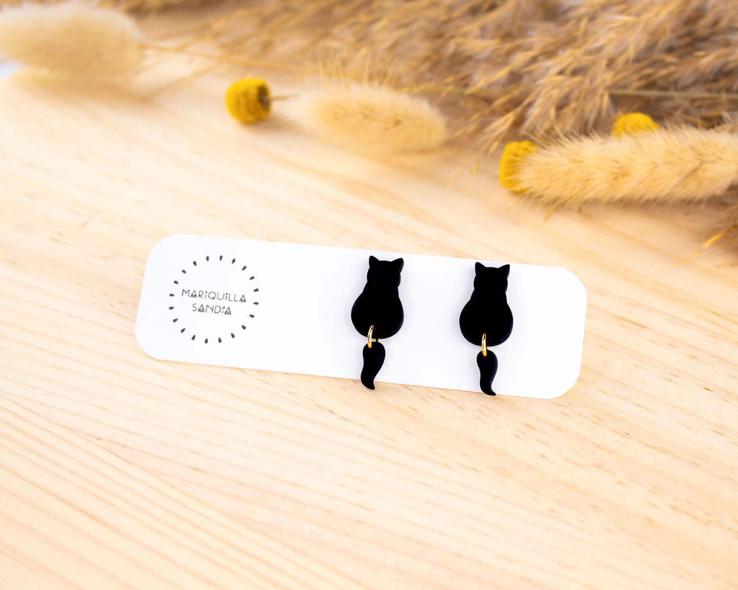 Black cat stud earrings with a dangling tail, handmade from hypoallergenic clay and displayed on a branded card.