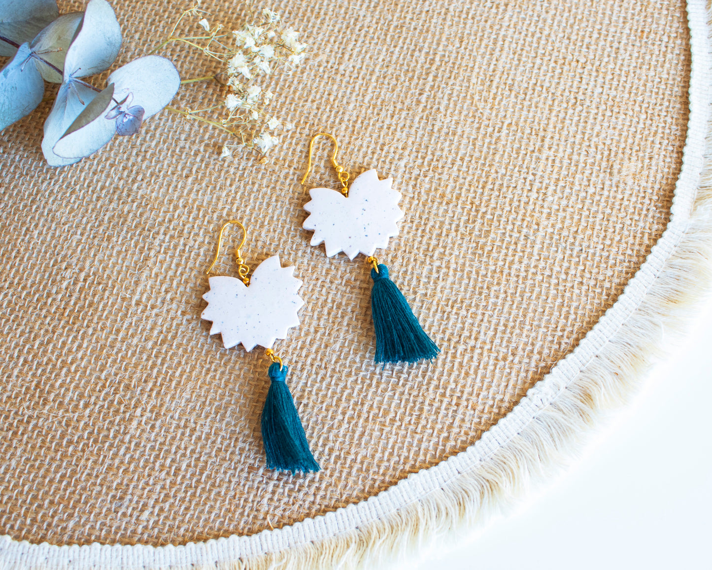 Close-up of handmade clay earrings with teal tassels on a woven background.
