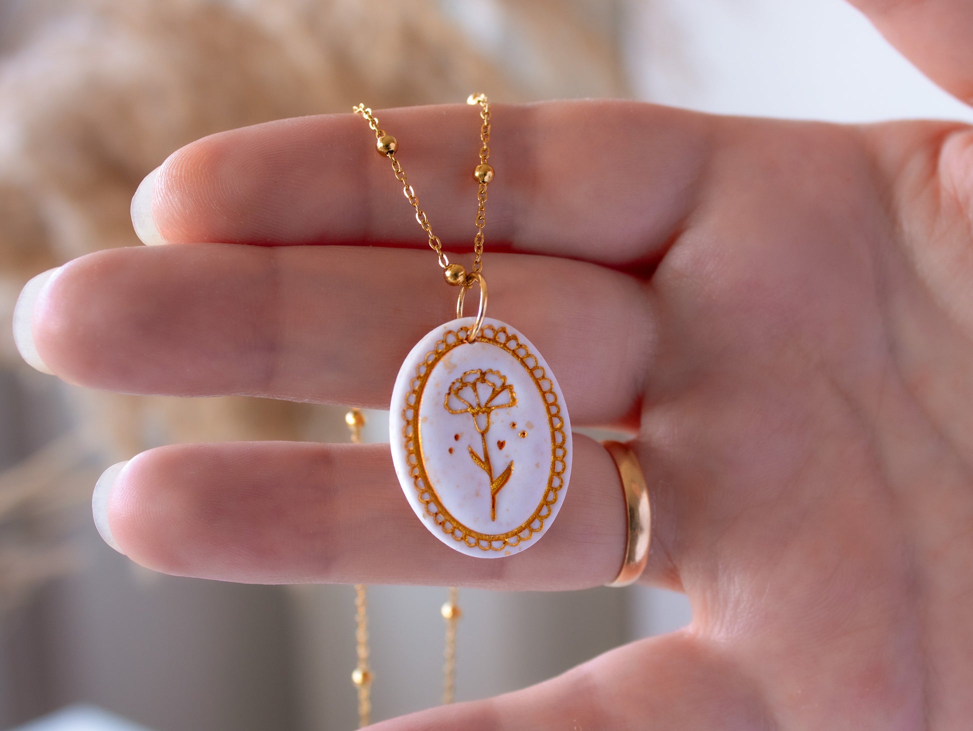 Close up of a hand holding a dainty birthflower necklace. The white clay pendant features a hand-painted bas-relief carnation for January in gold mica. It hangs from a hypoallergenic gold satellite chain.