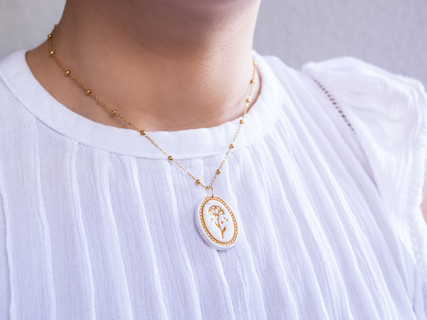 A woman wearing a white blouse models a custom birth month gift. She is wearing a hypoallergenic gold botanical pendant with a hand-painted birth flower design on a delicate gold satellite chain.