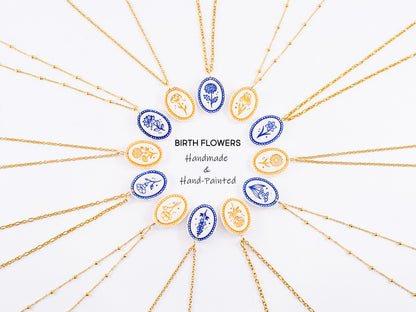 Twelve hand-painted birthflower necklaces arranged in a circle, alternating in gold mica and cobalt blue finishes. These hypoallergenic gold botanical pendants surround central text reading "Birth Flowers Handmade & Hand-Painted". A beautiful custom birth month charm collection and the perfect personalized jewelry for mom