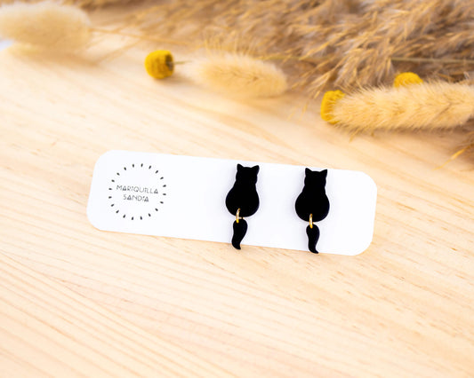 Black cat stud earrings with a dangling tail, handmade from hypoallergenic clay and displayed on a branded card.