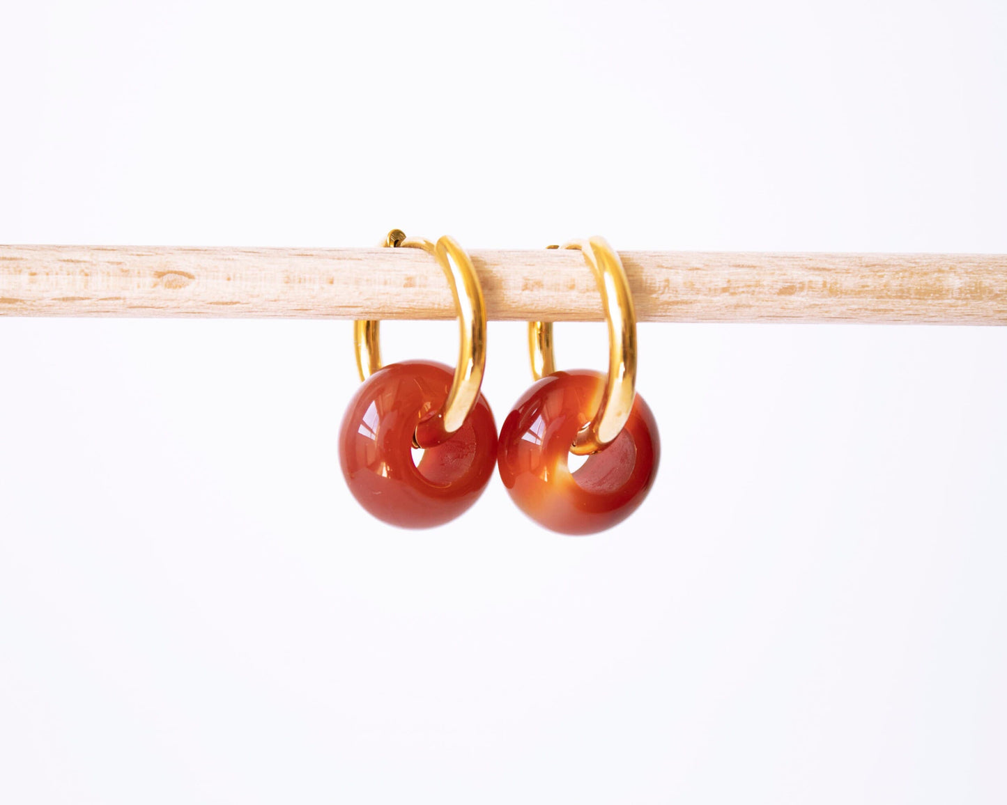 A pair of 18K gold plated hoop earrings with translucent Carnelian donut stone charms showing beautiful orange and red gradients, displayed on a wooden rod.