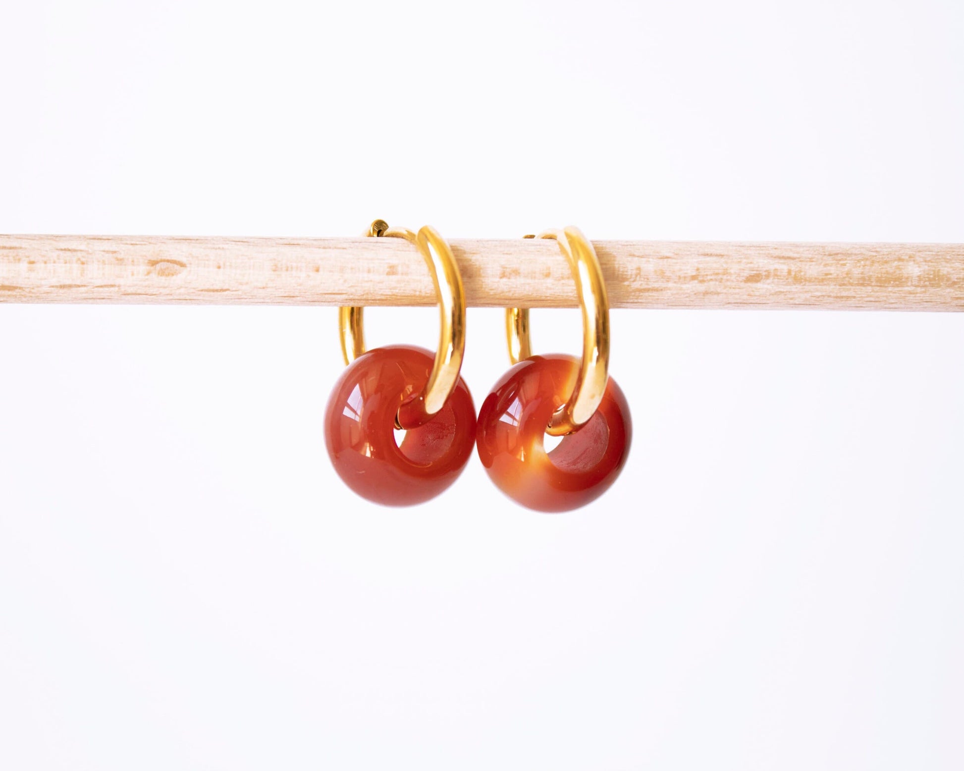 A pair of 18K gold plated hoop earrings with translucent Carnelian donut stone charms showing beautiful orange and red gradients, displayed on a wooden rod.