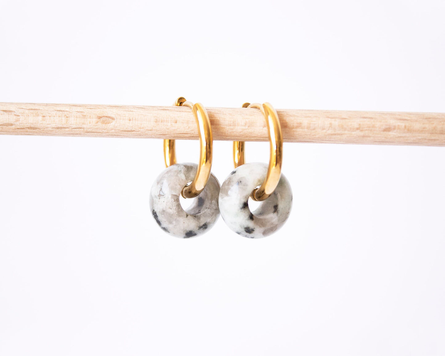 A pair of 18K gold plated hoop earrings featuring genuine Dalmatian Jasper donut stone charms with a unique speckled pattern, displayed on a wooden rod.