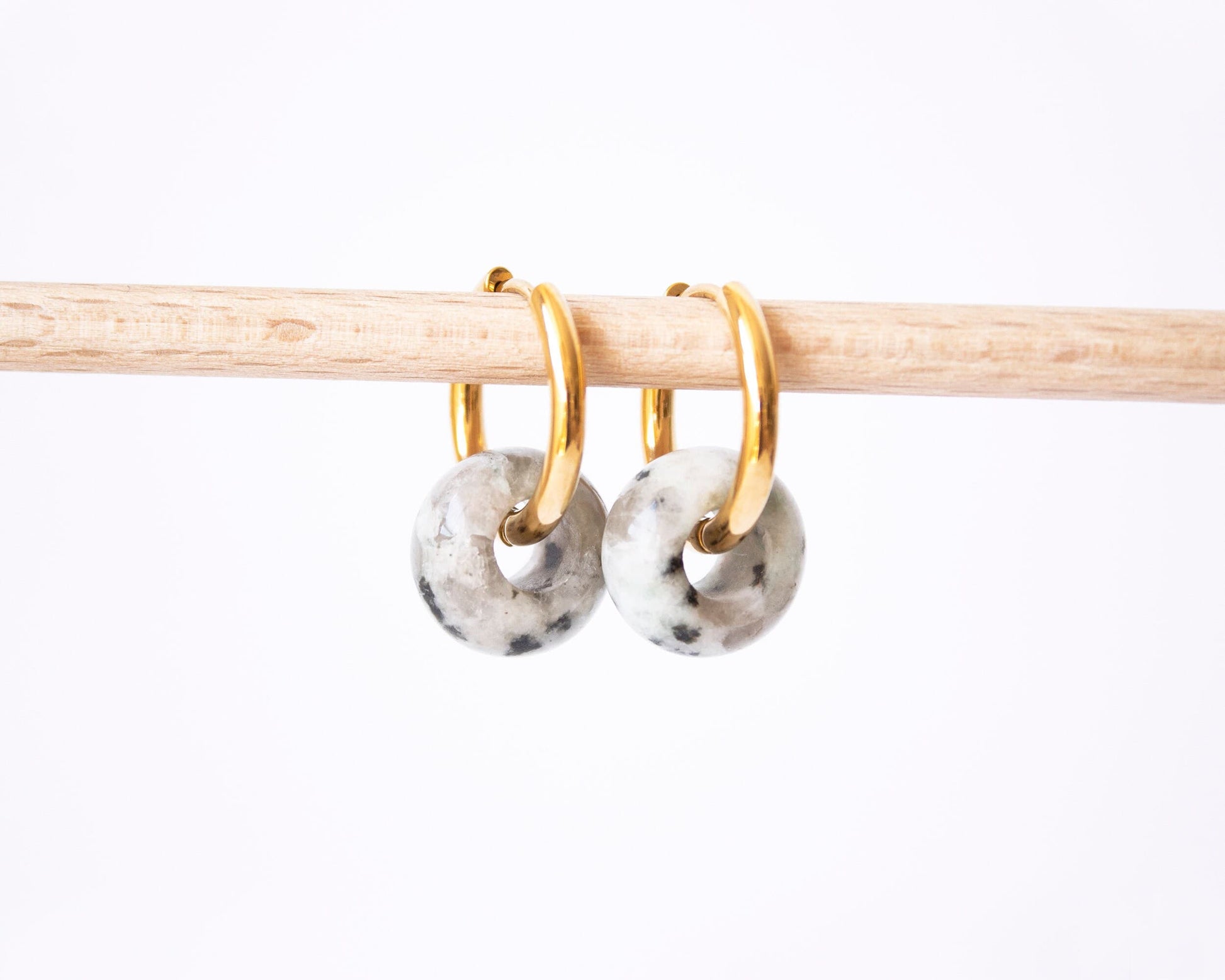 A pair of 18K gold plated hoop earrings featuring genuine Dalmatian Jasper donut stone charms with a unique speckled pattern, displayed on a wooden rod.