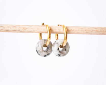A pair of 18K gold plated hoop earrings featuring genuine Dalmatian Jasper donut stone charms with a unique speckled pattern, displayed on a wooden rod.