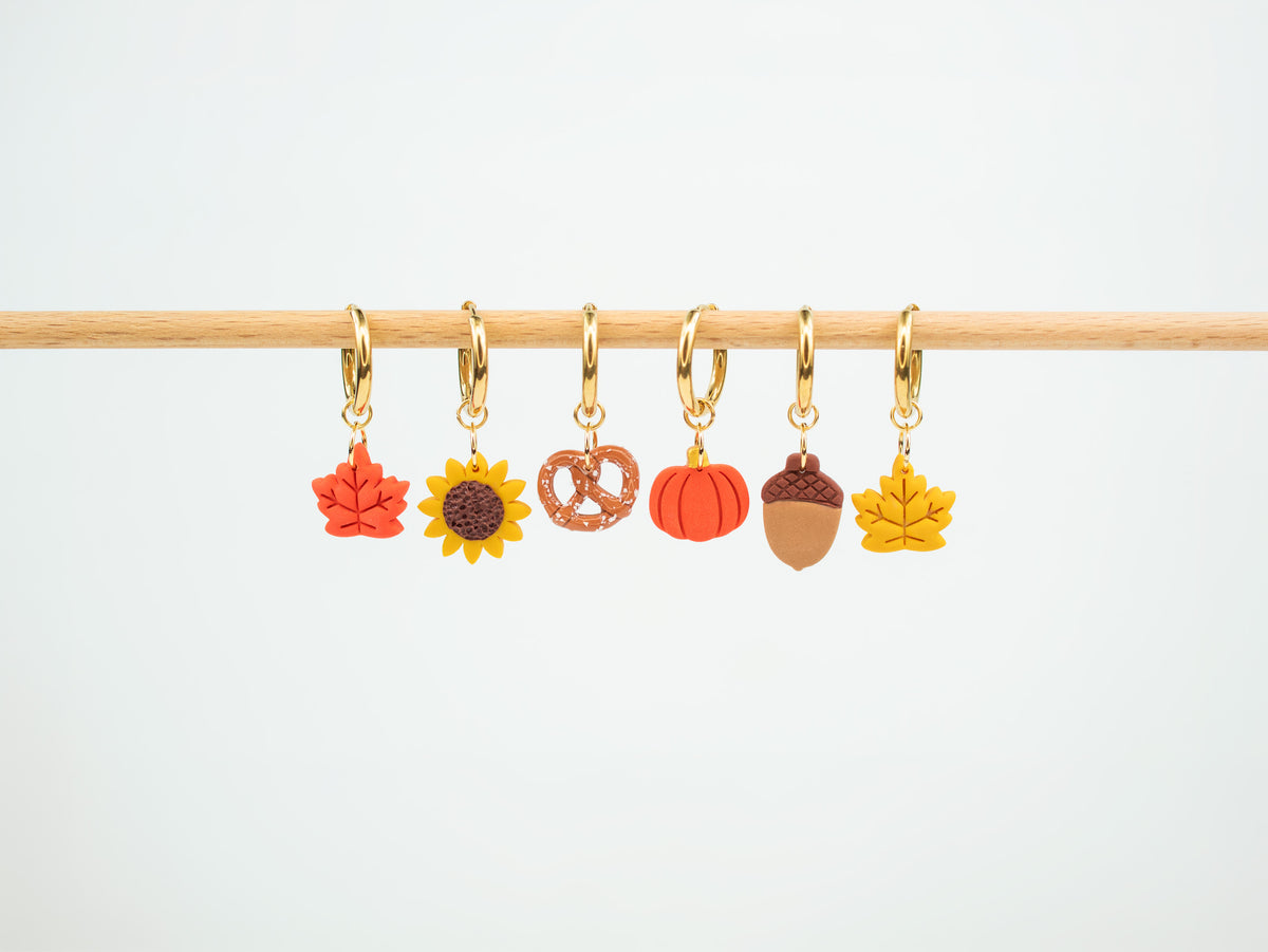 Collection of handmade autumn charm hoop earrings displaying interchangeable fall leaves, sunflower, pretzel, pumpkin, and acorn pendants.