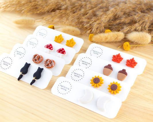 Handmade fall-themed stud earrings collection featuring designs of sunflowers, acorns, moons, black cats, mushrooms, pretzels, and colorful leaves