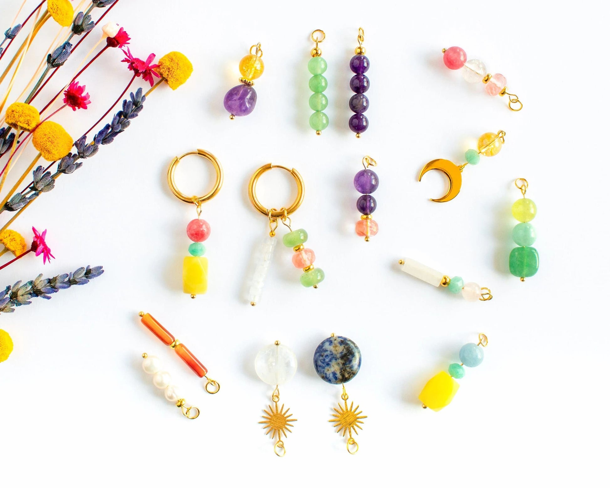 Gold hoop earrings next to a variety of interchangeable natural stone charms like amethyst and rose quartz on a white background with dried flowers.
