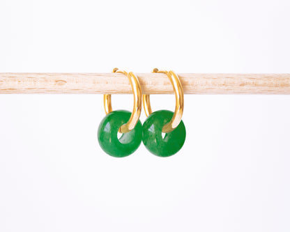 A pair of 18K gold plated hoop earrings with genuine Green Jade donut stone charms, displayed on a natural wooden rod.