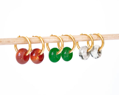 he complete collection of the interchangeable gemstone donut hoop earring set, with Carnelian, Green Jade, and Dalmatian Jasper charms shown side-by-side on a wooden display.