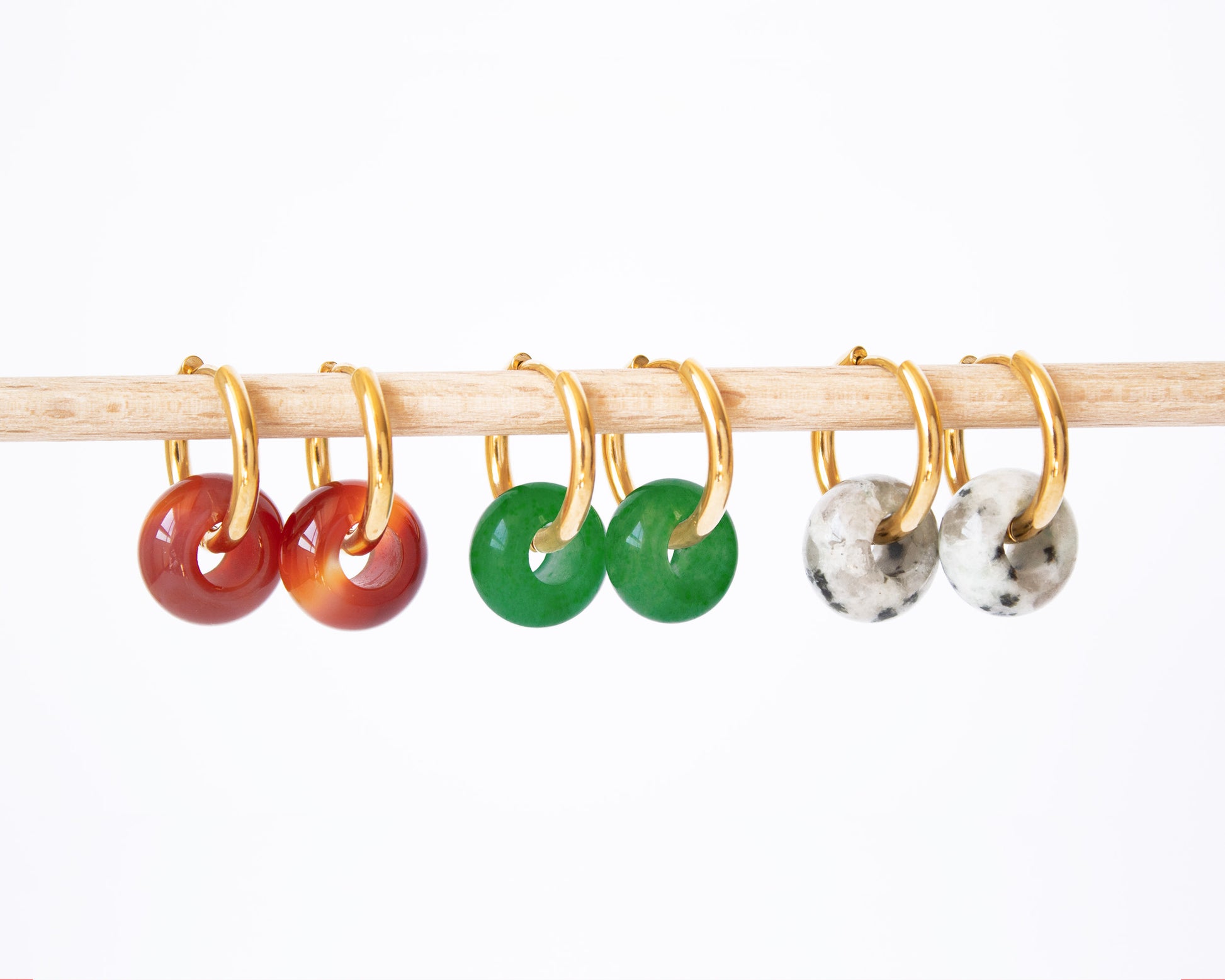 Interchangeable 18K gold plated hoop earrings displayed on a wooden rod, featuring all three natural stone donut charms: Carnelian, Green Jade, and speckled Dalmatian Jasper.