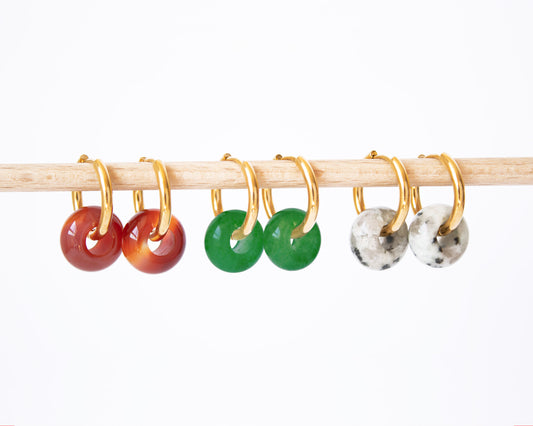 Interchangeable 18K gold plated hoop earrings displayed on a wooden rod, featuring all three natural stone donut charms: Carnelian, Green Jade, and speckled Dalmatian Jasper.